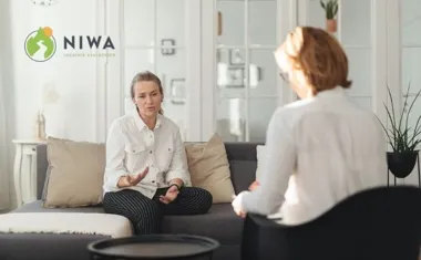 Dwie kobiety rozmawiają na kanapie w jasnym salonie, na ścianie logo NIWA.